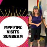 MPP Fife Visits Sunbeam! Photo of Sunbeam CEO Brian Swainson and MPP Catherine Fife with Sunbeam caps on standing in front of two flags.